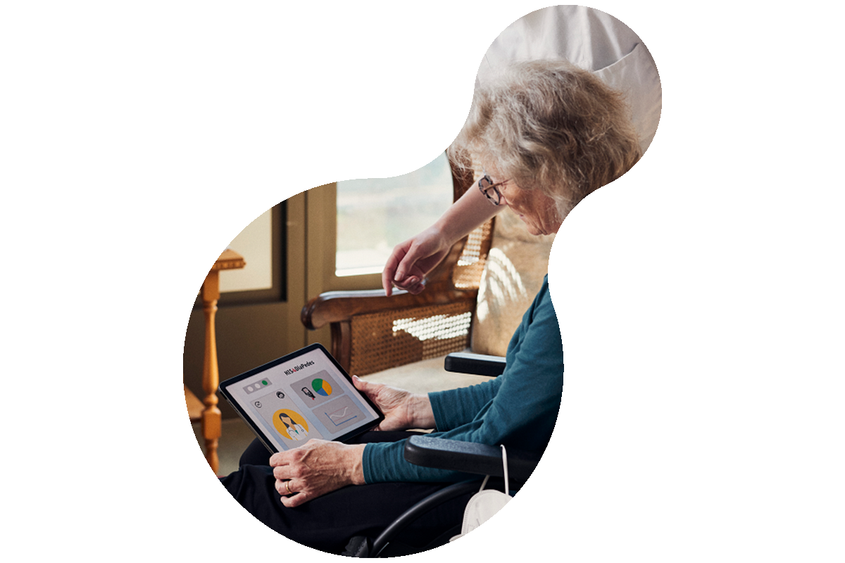 Foto einer älteren Frau im Rollstuhl, die ein Tablet hält. Neben ihr steht eine Pflegefachkraft, die ihr etwas erklärt.